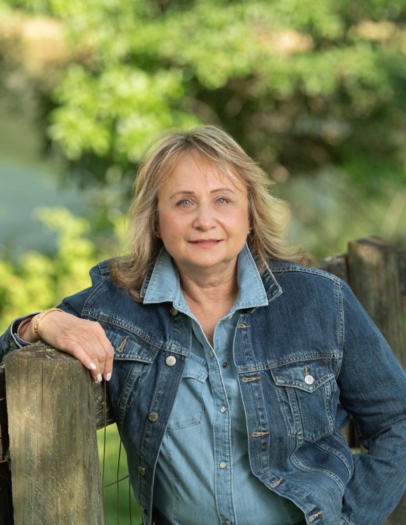 Photo of Susan S. Platt wearing a denim jacket leaning against a wood fence with trees in the background.